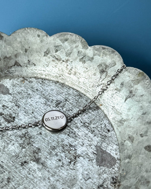 IBI NOTE Armband aus silberfarbenem Edelstahl mit "05.11.21" ♡ auf runder Platte graviert, auf Zinkschale vor blauem Hintergrund