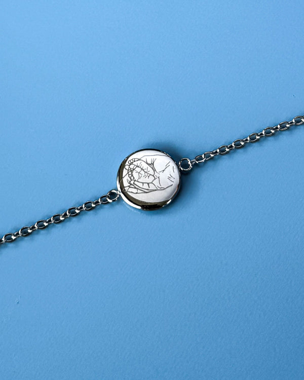 IBI PORTRAIT Armband aus silberfarbenem Edelstahl mit Strichzeichnung von Händen auf runder Platte graviert, vor blauem Hintergrund