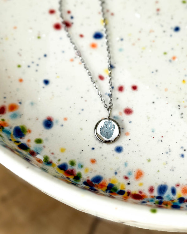 AYA MOTIF Halskette aus silberfarbenem Edelstahl mit kleinem Handabdruck auf rundem Anhänger graviert, auf gesprenkelter Keramikschale