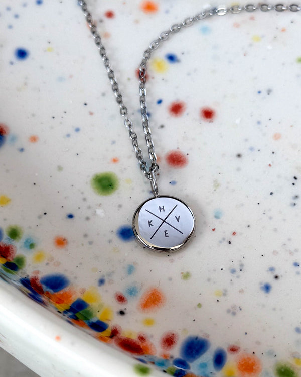 AYA X Halskette aus silberfarbenem Edelstahl mit den Initialen "H K V E" kreuzförmig auf kleinem Anhänger graviert, auf gesprenkelter Keramikschale