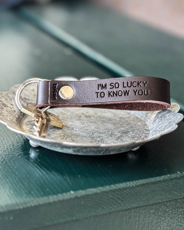 BOND Schlüsselanhänger aus dunkelbraunem Leder mit der Gravur "I'm so lucky to know you" und Schlüssel in Zinkschale auf grünem Tisch