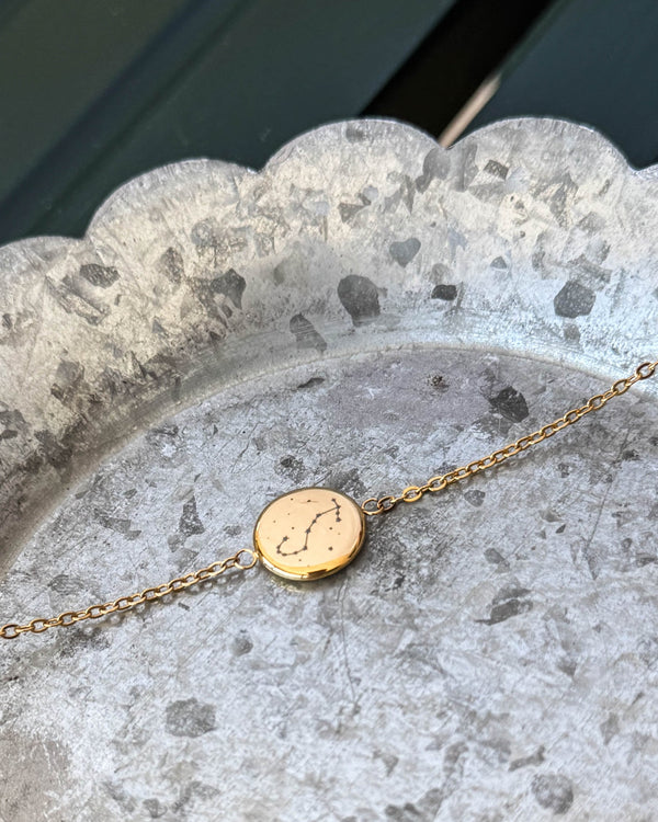 IBI STAR Armband aus vergoldetem Edelstahl mit dem Sternbild Skorpion auf runder Platte graviert, auf Zinkschale liegend