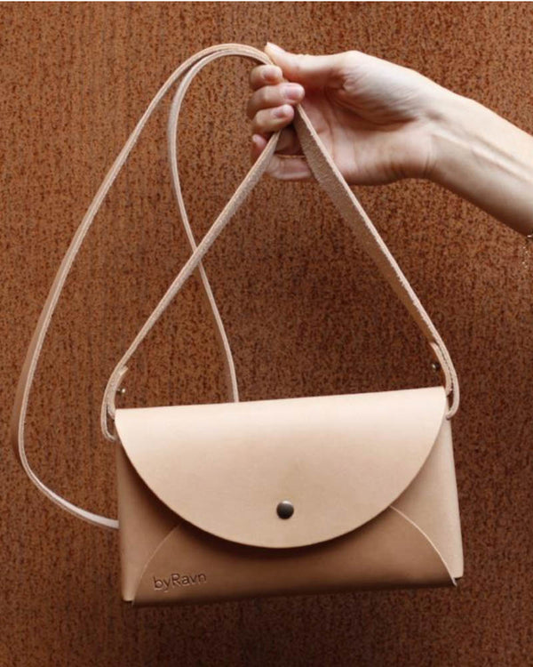 INA Schultertasche aus naturfarbenem Leder, am Riemen hochgehalten vor rostroter Wand