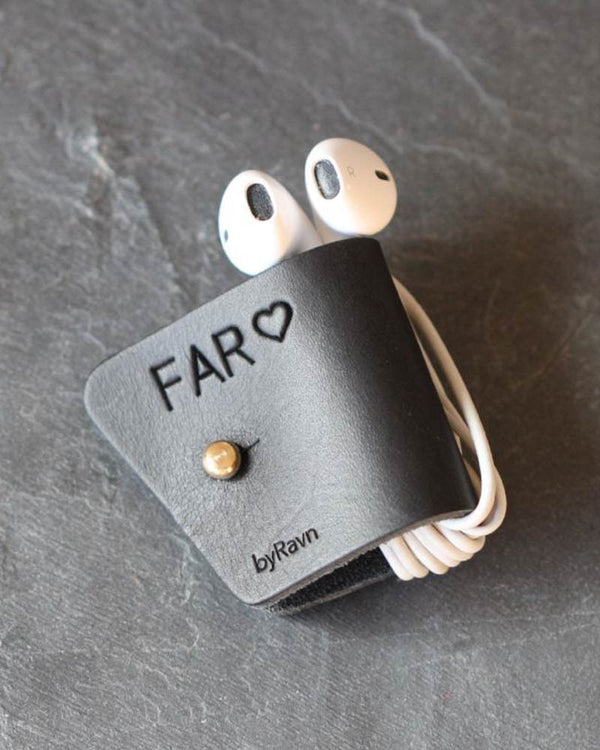 KAX Kopfhörerhalter aus schwarzem Leder mit persönlicher Gravur "Far" ♡ und Messingknopf, mit Earpods auf dunklem Schiefer