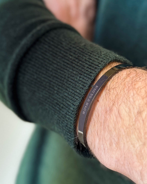 MARS SCRIPT Armband aus schwarzem Edelstahl mit den Daten "18.04.16 ♡ 02.09.18" in Handschrift graviert, zu dunkelgrünem Strick getragen