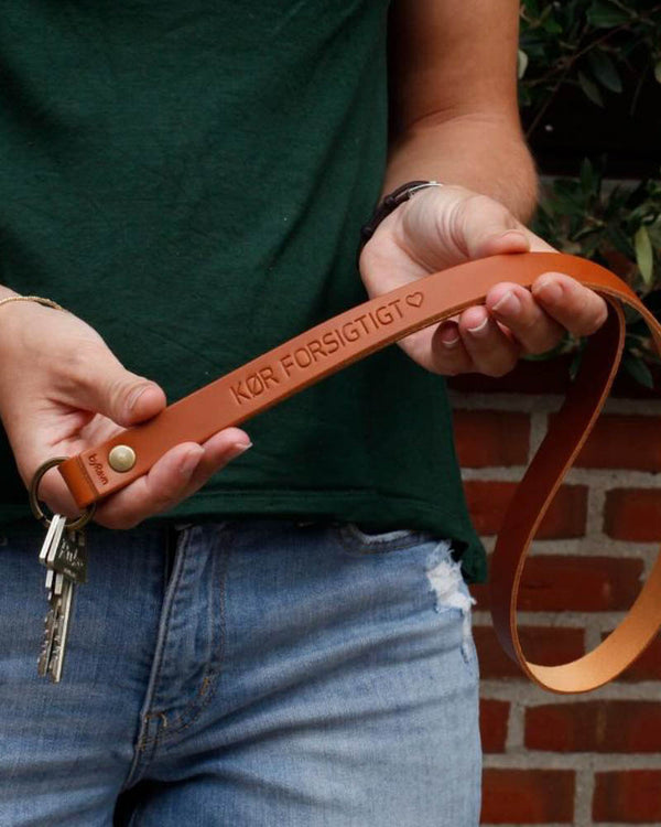 MAX Schlüsselanhänger aus Cognac-Leder als lange Schlaufe mit der Gravur "Kør forsigtigt" ♡, in den Händen gehalten mit Schlüsseln