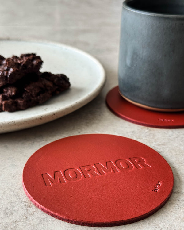 MOKKA Untersetzer aus rotem Leder mit Prägung "Mormor" auf hellem Tisch mit grauer Kaffeetasse und Teller mit Schokoladenkuchen