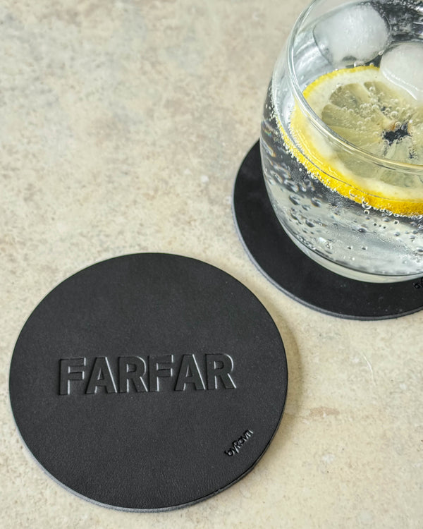 MOKKA Untersetzer aus schwarzem Leder mit Prägung "Farfar" auf hellem Tisch neben Glas mit Gin Tonic und Zitronenscheibe