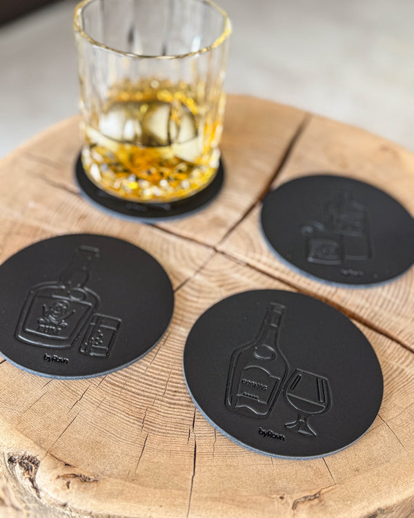 MOKKA Spirituosen-Untersetzer aus schwarzem Leder mit Prägung Rum, Cognac und Gin auf Holzscheibe neben Kristallglas mit Whisky