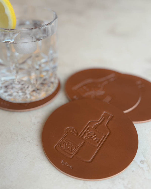 MOKKA Spirituosen-Untersetzer aus cognacfarbenem Leder mit geprägter Ginflasche und Drink, auf heller Steinfläche neben Glas mit Zitronenscheibe
