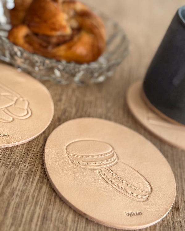 MOKKA Kaffee & Kuchen-Untersetzer aus naturfarbenem Leder mit geprägten Macarons auf Holztisch neben Zimtschnecke und schwarzer Tasse