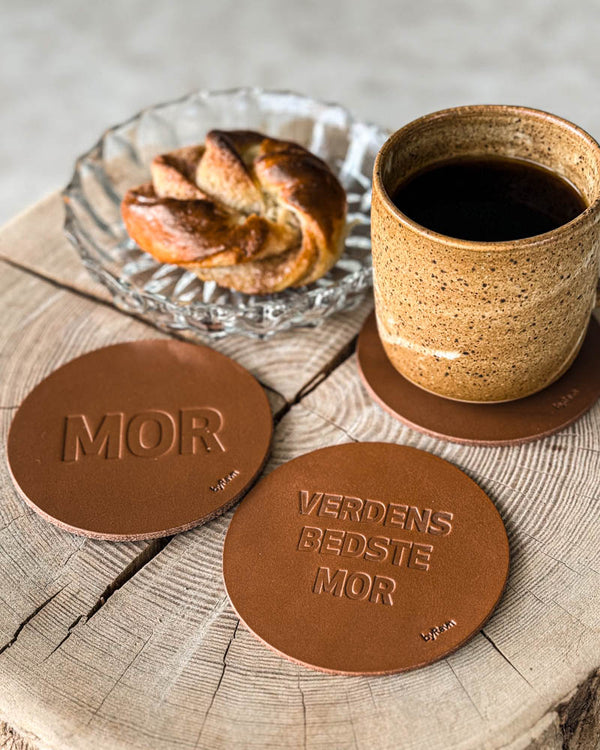 MOKKA Untersetzer aus cognacfarbenem Leder mit Prägung "Mor" und "Verdens bedste mor" auf Holzscheibe mit Kaffeetasse und Zimtschnecke