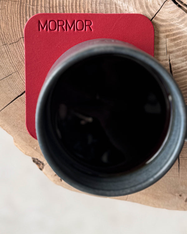 MOKKA quadratischer Untersetzer aus rotem Leder mit der Gravur "Mormor" unter schwarzer Kaffeetasse auf Holzscheibe, von oben