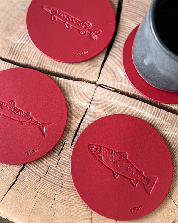 MOKKA Angler-Untersetzer aus rotem Leder mit geprägter Meerforelle, Makrele und Hecht auf Holzscheibe neben grauer Kaffeetasse