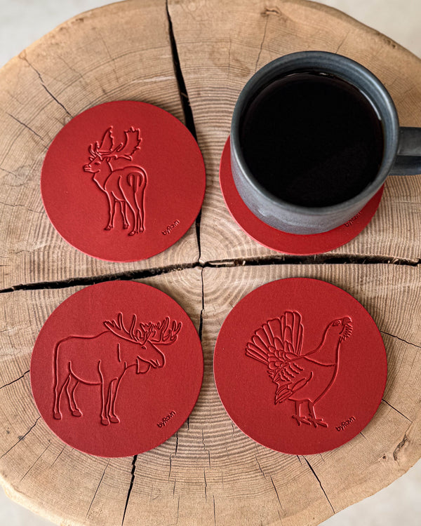MOKKA Jagd-Untersetzer aus rotem Leder mit geprägtem Elch, Auerhahn und Damhirsch auf Holzscheibe neben grauer Kaffeetasse auf Untersetzer