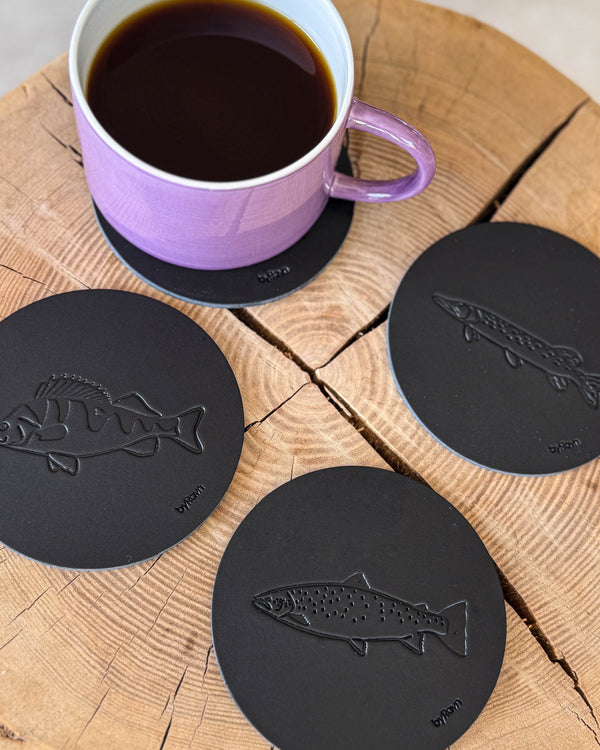 MOKKA Angler-Untersetzer aus schwarzem Leder mit geprägter Meerforelle, Barsch und Hecht auf Holzscheibe neben lila Kaffeetasse