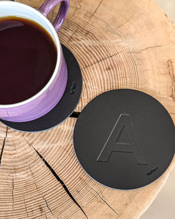 MOKKA Untersetzer aus schwarzem Leder mit geprägtem Buchstaben "A" auf Holzscheibe neben lila Kaffeetasse auf Untersetzer