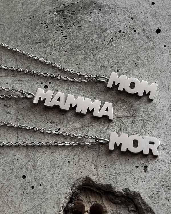 MOM Halsketten aus silberfarbenem Edelstahl mit Buchstabenanhängern "MOM", "MAMMA" und "MOR", liegend auf rohem Beton