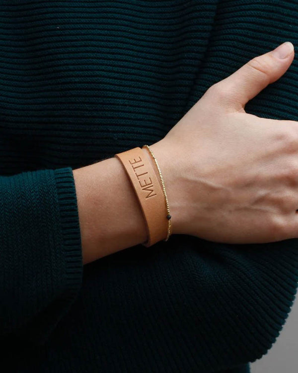 MONO Lederarmband aus naturfarbenem Leder mit graviertem Namen "Mette" am Handgelenk, kombiniert mit zartem goldfarbenem Armband