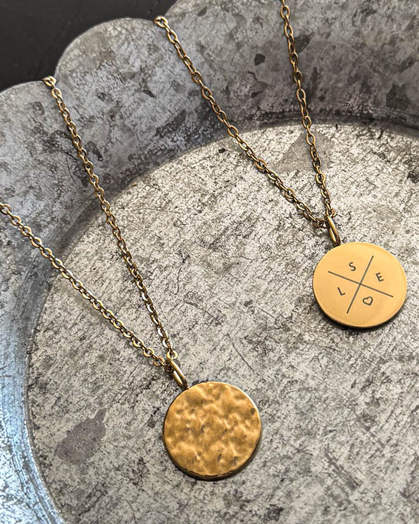 MOON X Halskette aus vergoldetem Edelstahl mit Kreuz und Initialen "S E L" und Herz graviert, gehämmerte Münze daneben, auf Zinkschale