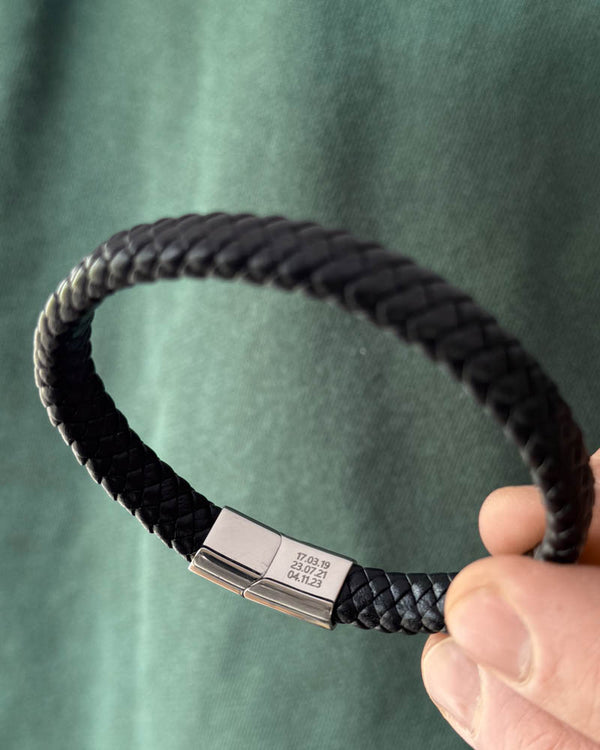 RAY Lederarmband aus schwarzem geflochtenem Leder in der Hand gehalten, die Daten "17.03.19, 23.07.21, 04.11.23" auf dem Verschluss graviert