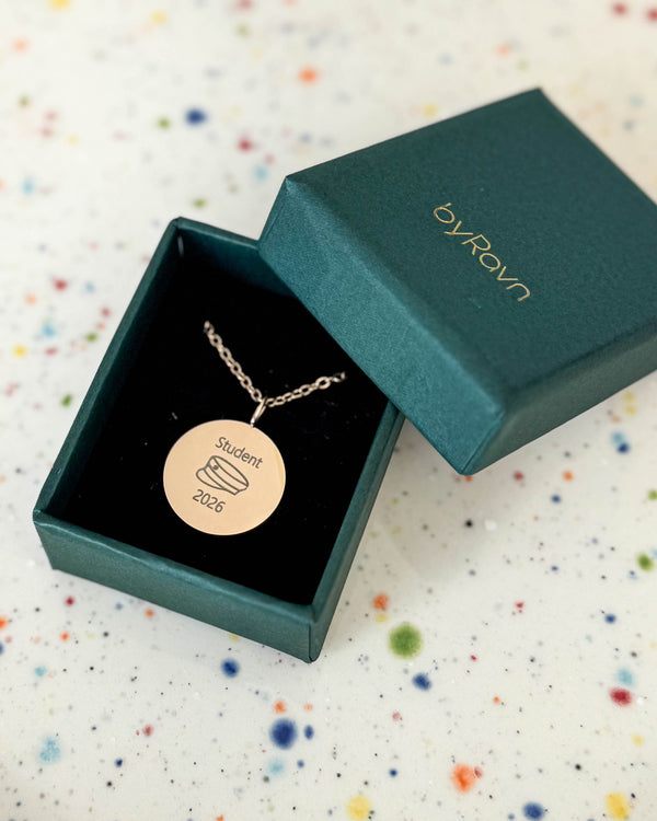 SIA MOMENTS Halskette aus vergoldetem Edelstahl mit Studentenmütze und "Student 2026" graviert, in grüner byRavn Geschenkbox auf gesprenkelter Schale