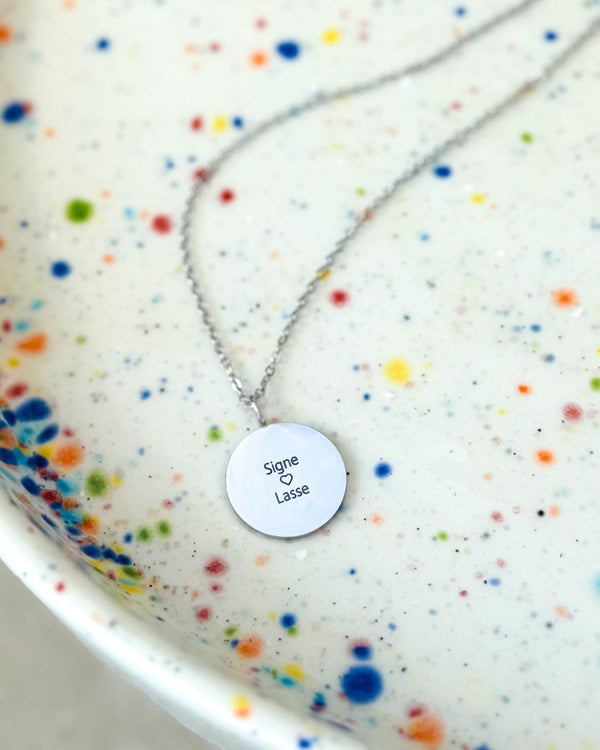 SIA QUOTE Namenskette aus silberfarbenem Edelstahl mit der Gravur "Signe ♡ Lasse" auf rundem Anhänger, auf gesprenkelter Schale