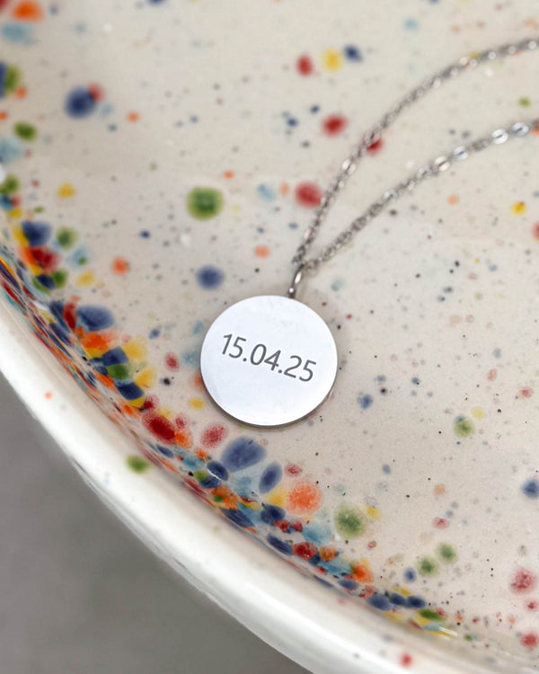SIA NOTE Halskette aus silberfarbenem Edelstahl mit dem Datum "15.04.25" auf rundem Anhänger graviert, auf gesprenkelter Keramikschale