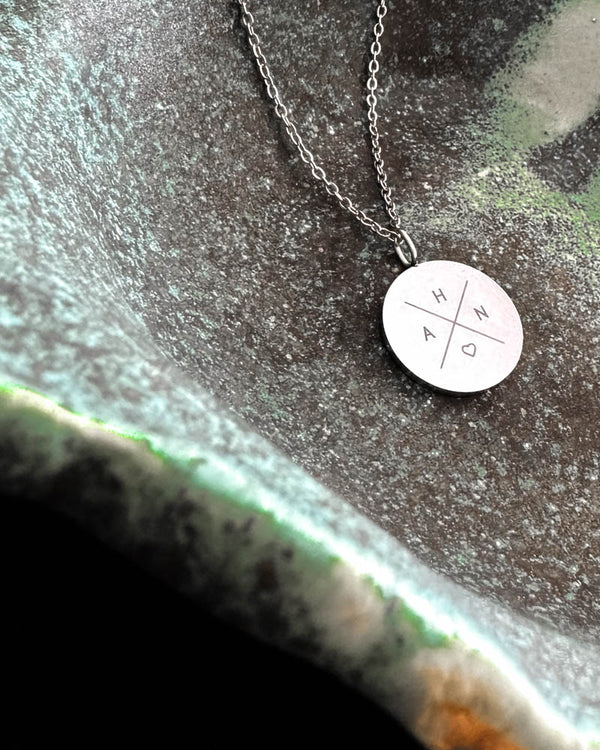 SIA X Halskette aus silberfarbenem Edelstahl mit den Initialen "H A N ♡" kreuzförmig auf rundem Anhänger graviert, in grüner Keramikschale
