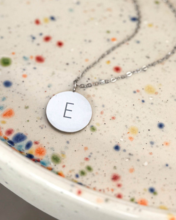 SIA LETTER Halskette aus silberfarbenem Edelstahl mit dem Buchstaben "E" auf rundem Anhänger graviert, auf gesprenkelter Keramikschale