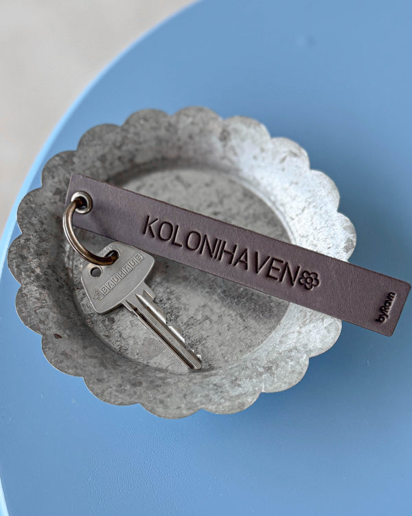 SOLO Large Schlüsselanhänger aus dunkelbraunem Leder mit der Gravur "Kolonihaven" und Blume, mit Schlüssel in Zinkschale
