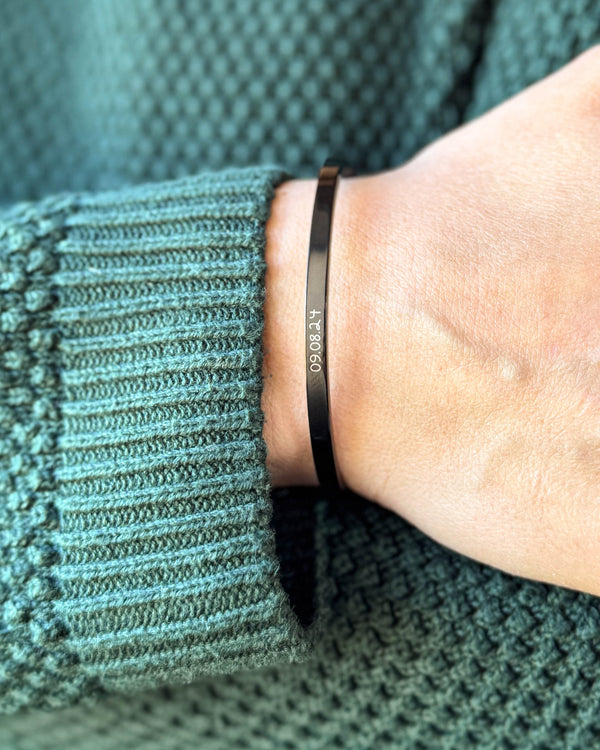 VENUS SCRIPT Armband aus schwarzem Edelstahl mit dem Datum "09.08.24" in Handschrift am Handgelenk, grüner Strickpullover