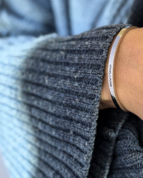 VENUS Armband aus silberfarbenem Edelstahl mit der Gravur "Maria ♡ Simon" am Handgelenk, blauer Strickpullover