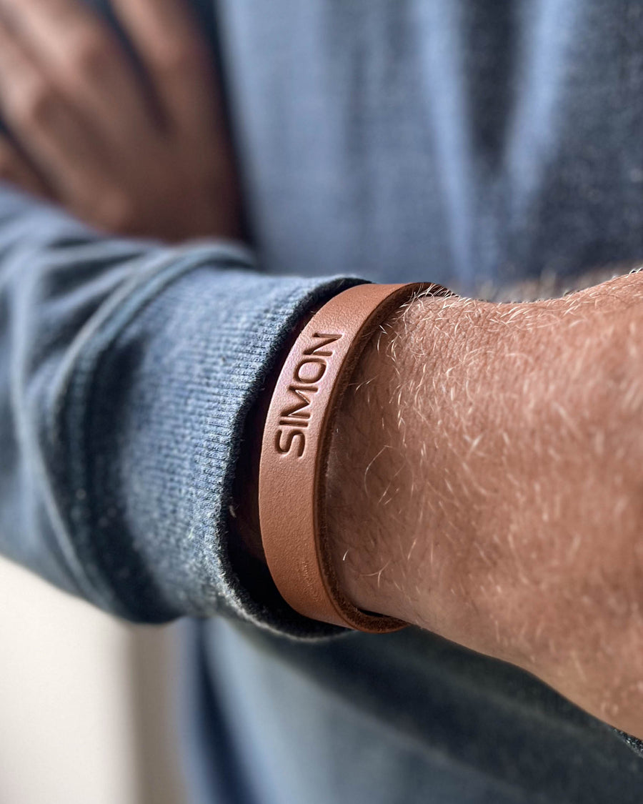 MONO Lederarmband in Cognac mit graviertem Namen "Simon" am Handgelenk eines Mannes in blauem Strickpullover