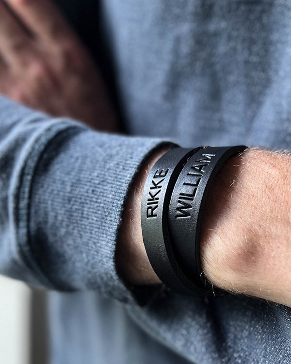 DUO Lederarmband in Schwarz mit gravierten Namen "Rikke" und "William" am Handgelenk eines Mannes in grauem Strickpullover