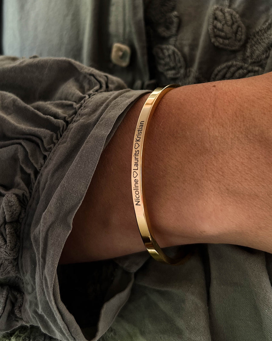 VENUS Armband aus vergoldetem Edelstahl mit der Gravur "Nicoline ♡ Laurits ♡ Kristian" am Handgelenk, graue Bluse