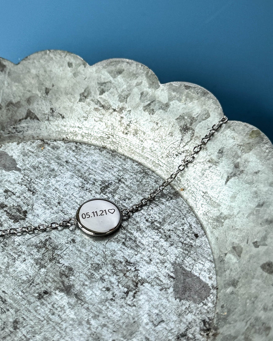 IBI NOTE Armband aus silberfarbenem Edelstahl mit "05.11.21" ♡ auf runder Platte graviert, auf Zinkschale vor blauem Hintergrund