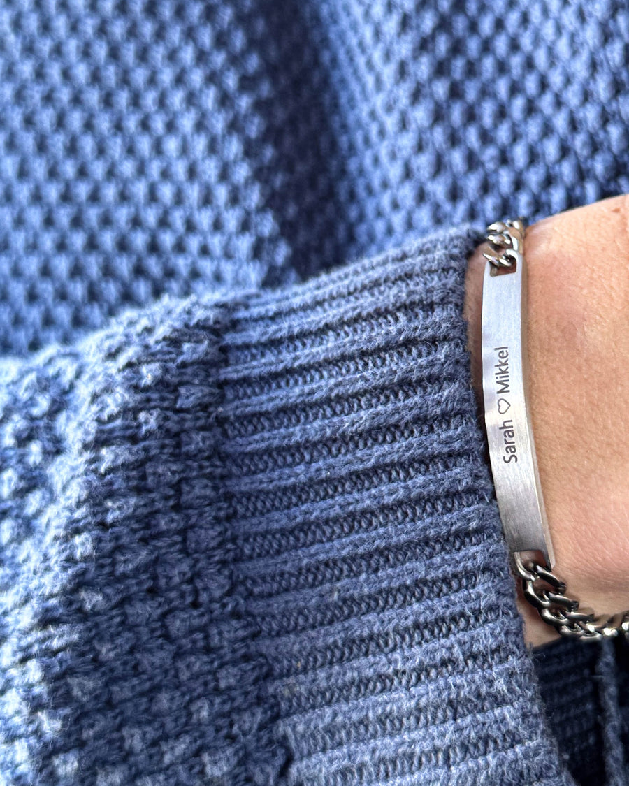 AVA Armband aus gebürstetem silberfarbenem Edelstahl mit Gravur "Sarah ♡ Mikkel" am Handgelenk, blauer Strickpullover