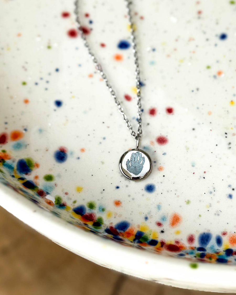 AYA MOTIF Halskette aus silberfarbenem Edelstahl mit kleinem Handabdruck auf rundem Anhänger graviert, auf gesprenkelter Keramikschale