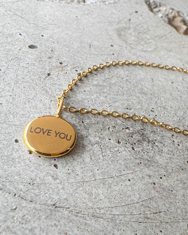 AYA NOTE Halskette aus vergoldetem Edelstahl mit der Gravur "Love you" auf kleinem rundem Anhänger, auf Beton liegend