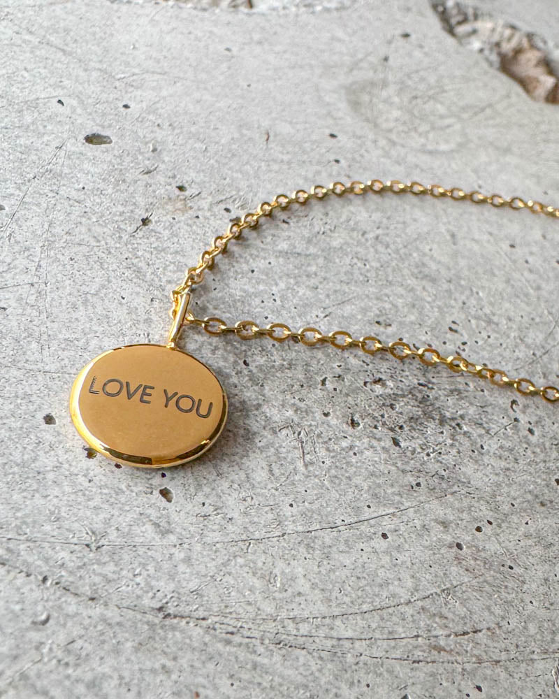 AYA NOTE Halskette aus vergoldetem Edelstahl mit der Gravur "Love you" auf kleinem rundem Anhänger, auf Beton liegend