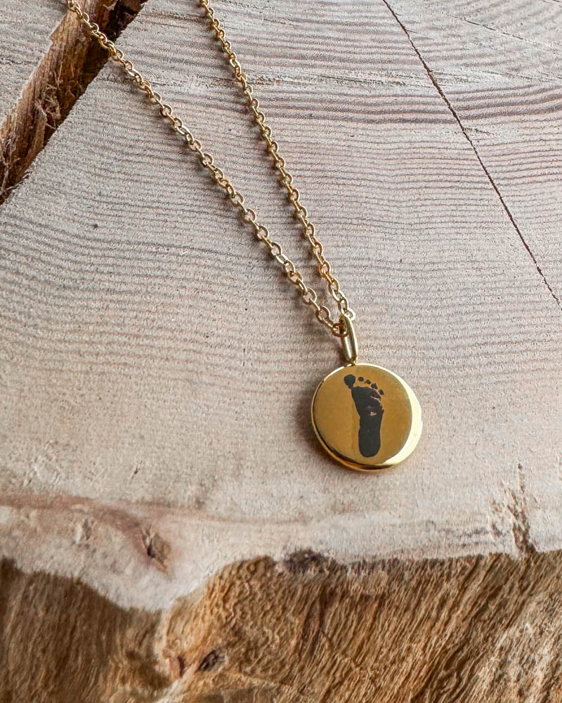 AYA MOTIF Halskette aus vergoldetem Edelstahl mit Babyfußabdruck auf kleinem Anhänger graviert, auf hellem Holz mit Riss