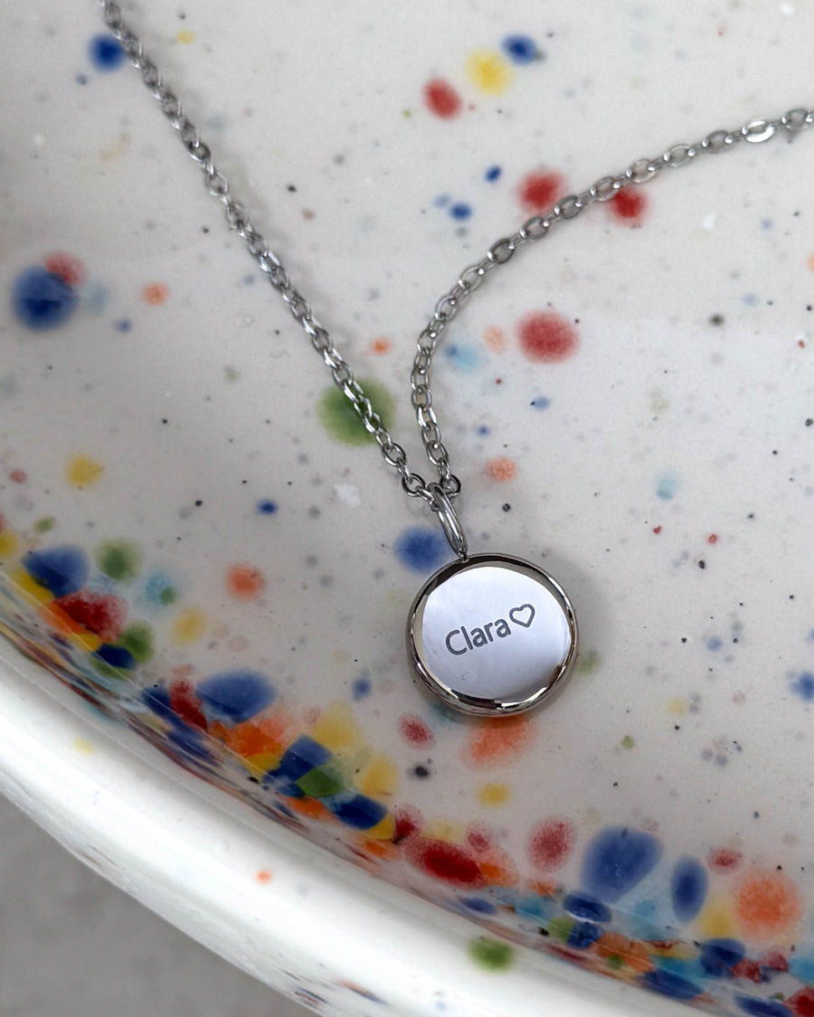AYA NOTE Namenskette aus silberfarbenem Edelstahl mit dem Namen "Clara" ♡ auf kleinem Anhänger graviert, auf gesprenkelter Keramikschale