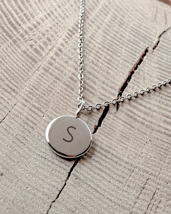 AYA LETTER Halskette aus silberfarbenem Edelstahl mit dem Buchstaben "S" auf kleinem rundem Anhänger graviert, auf hellem Holz mit Riss