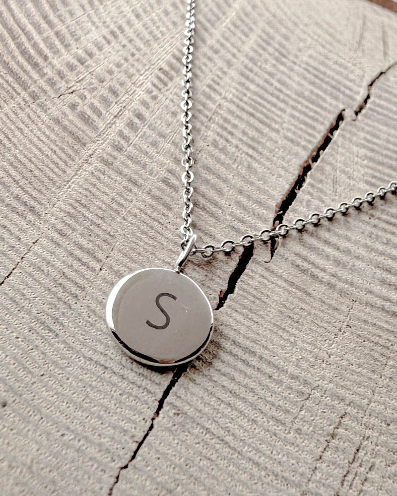 AYA LETTER Halskette aus silberfarbenem Edelstahl mit dem Buchstaben "S" auf kleinem rundem Anhänger graviert, auf hellem Holz mit Riss