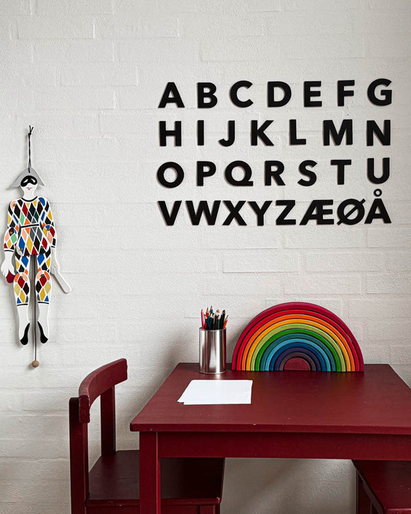 BOX Buchstaben aus schwarzem Leder als ganzes Alphabet A–Å an weißer Backsteinwand über rotem Kindertisch mit Holz-Regenbogen