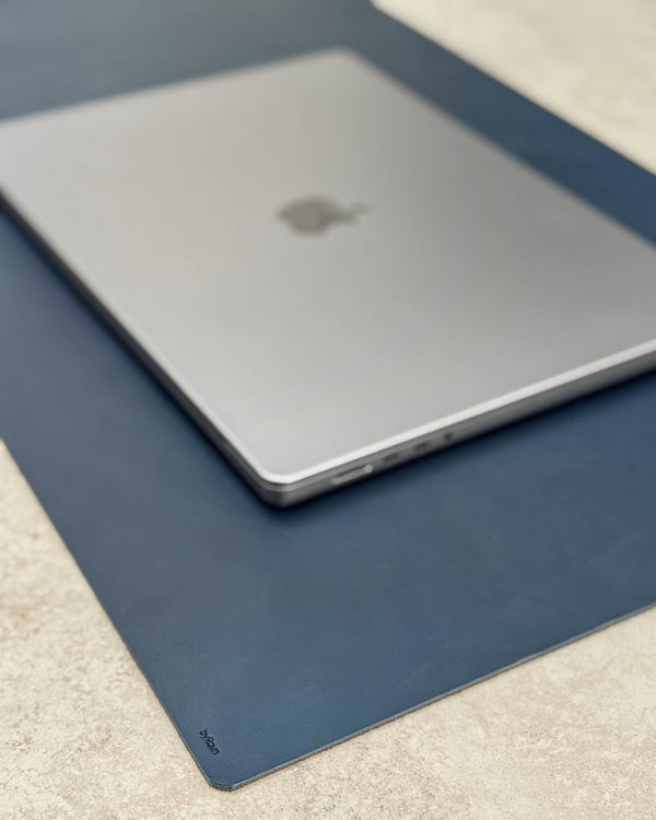 DESK Schreibtischunterlage aus dunkelblauem Leder mit byRavn-Prägung unter silberfarbenem Laptop auf heller Tischplatte