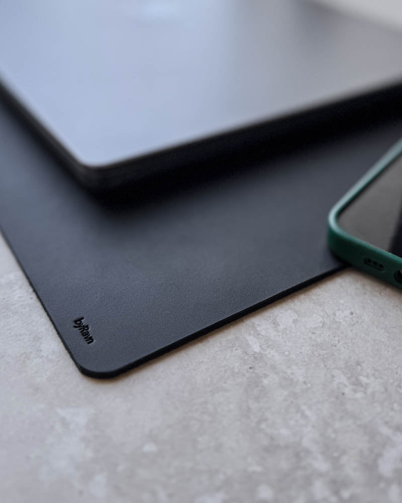 DESK Schreibtischunterlage aus schwarzem Leder, Nahaufnahme der Ecke mit byRavn-Prägung unter Laptop und Telefon