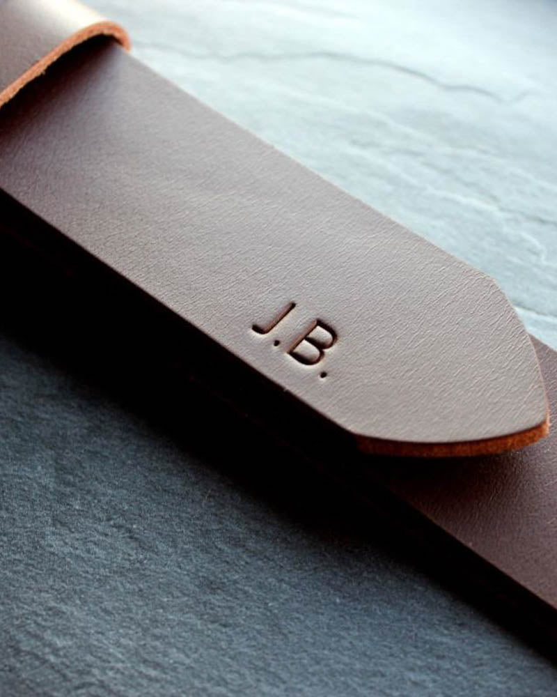EDGE Gürtel aus dunkelbraunem Leder in Nahaufnahme mit den Initialen "J.B." an der Spitze graviert, auf Schieferplatte