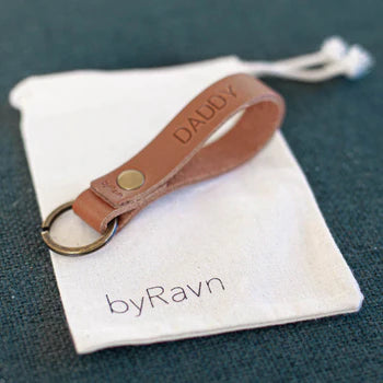 Geschenkbeutel aus weißem Stoff mit byRavn-Logo und Schlüsselanhänger aus Leder in Cognac mit der Gravur "Daddy" auf grünem Untergrund